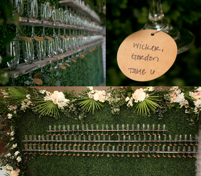 Champagne wall and table assignment.  Large background covered in greenery and topped with flowers.  Champagne flutes hanging with table assignment tags.